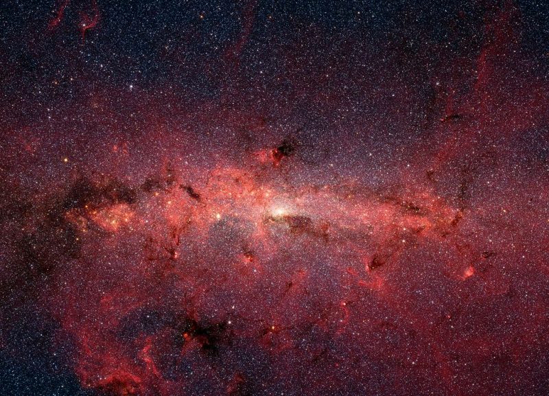 Droga Mleczna na zdjęciu w podczerwieni wykonanym przez Kosmiczny Teleskop Spitzera. Starsze chłodne gwiazdy są tu widoczne jako niebieskie, pył oświetlony przez duże gorące gwiazdy ma czerwonawy odcień, a duża biała plama pośrodku oznacza położenie Sagittariusa A, który jest uznawany za supermasywną czarną dziurę znajdującą się w centrum naszej galaktyki. Fot. NASA/JPL-Caltech/S. Stolovy (SSC/Caltech) 